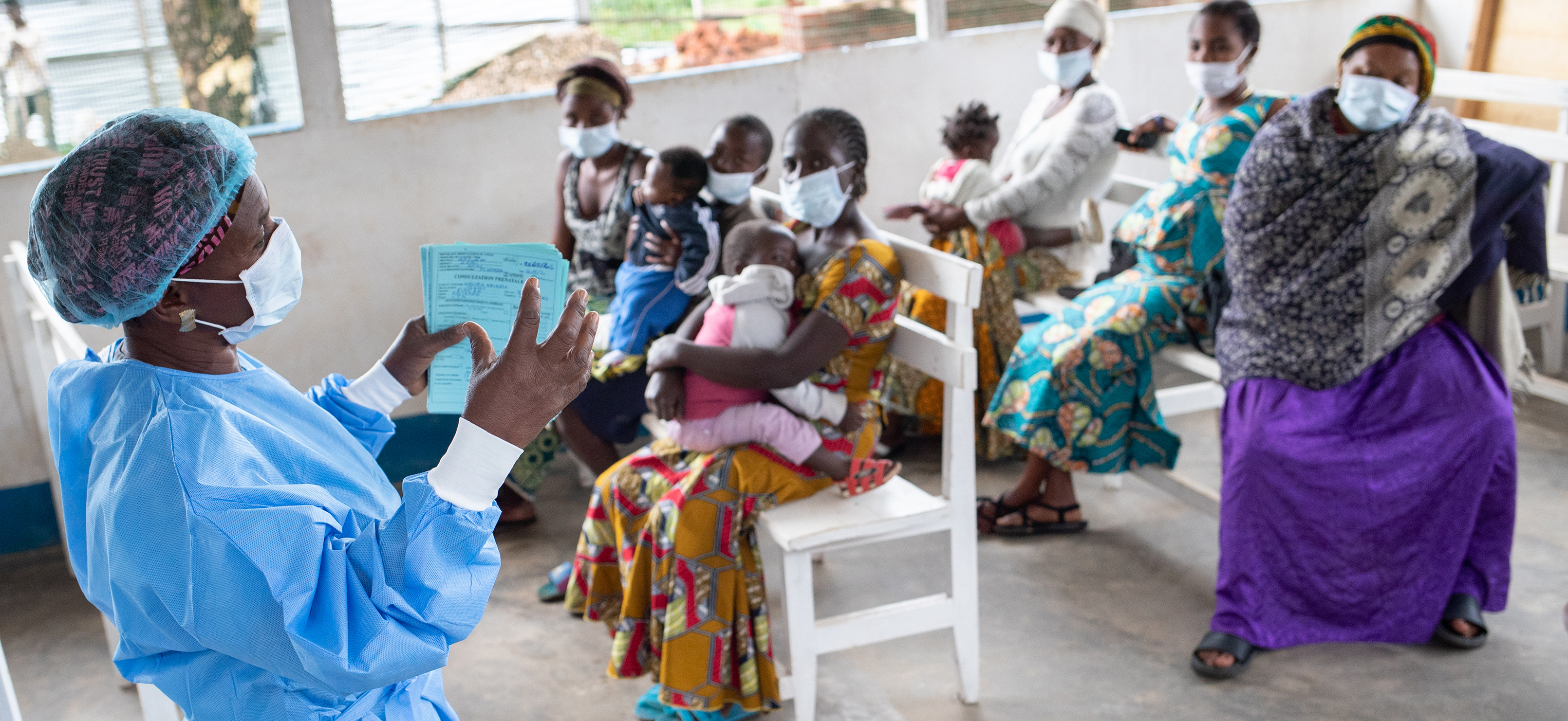 A Congolese nurse with a mask and blue scrubs presents outdoors in front of a group of seated women who are in masks and many have infants on their laps