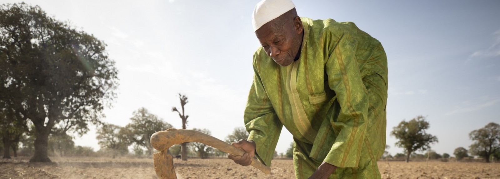Man in white cap and long green tunic tills dry and dusty land