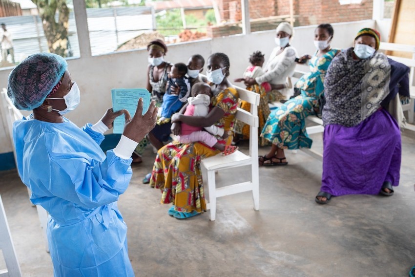 A nurse in blue protective equipment and white face mask speaks to a group of women with children seated in a courtyard. 