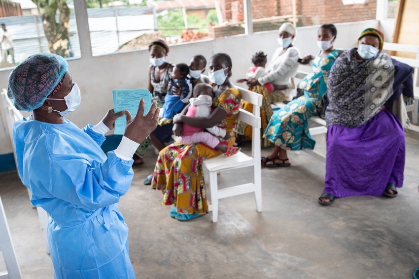 A nurse in blue protective equipment and white face mask speaks to a group of women with children seated in a courtyard.