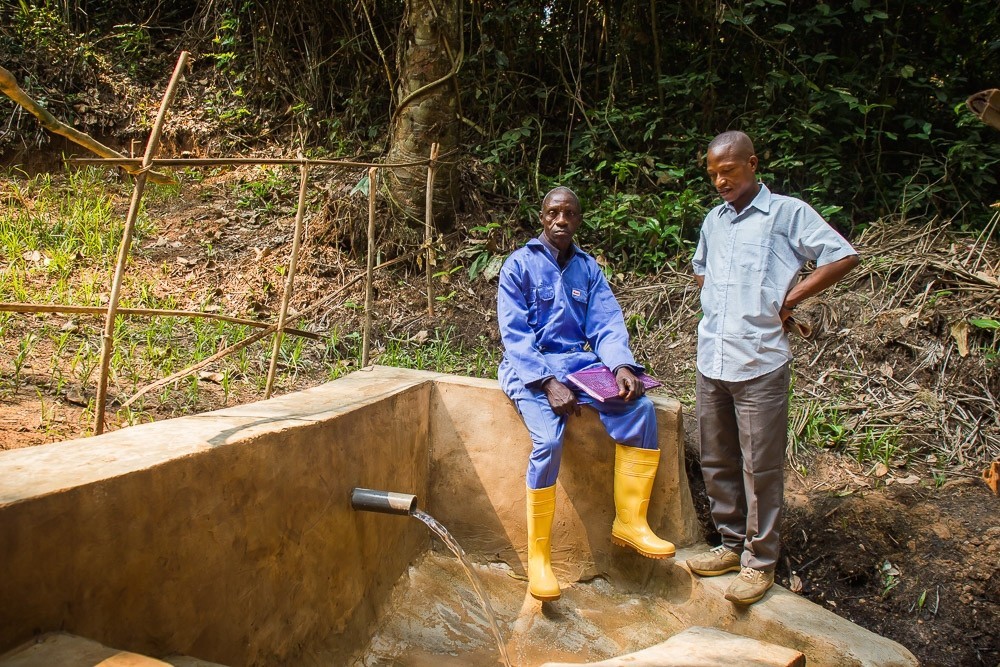 Two Congolese men, one seated and one standing, pose next to a water source with green vegetation behind them