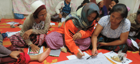 A group of women sit on a bright orange blanket writing on a large sheet of paper together