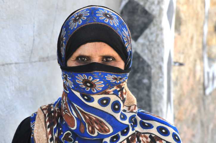 A Yemeni woman with a printed blue niqab stands in front of a gray wall and looks directly into a camera