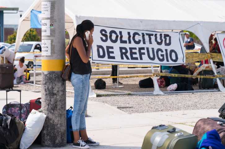 Woman stands next to a lamp post talking on her cell phone surrounded by luggage and belongings of fellow migrants at the border crossing